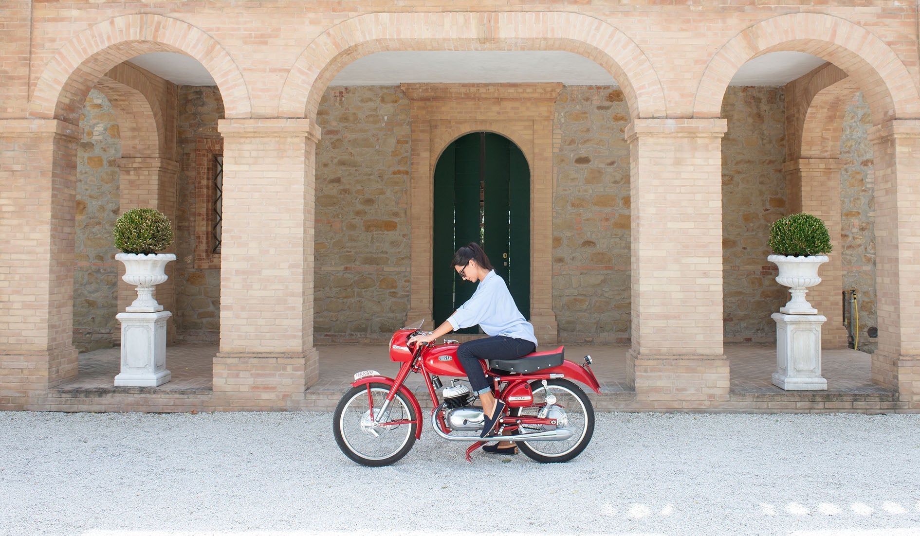 Luxury Denim Jeans handcrafted and made in Italy from Selvedge denim, woman sitting on antique red Ferrari motorcycle in front of Italian Villa