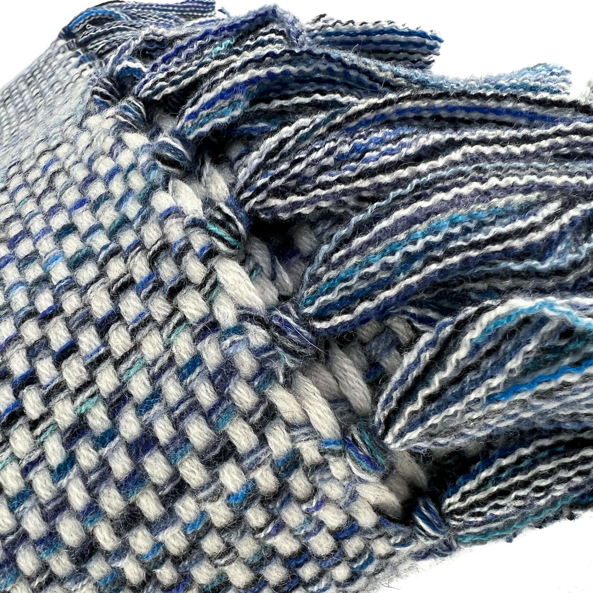 Close-up of an An Italian made Cashmere throw with a white weft and different shades of blue warp, folded on a white background, with hand knotted multiple shades of blue tassels showing in front