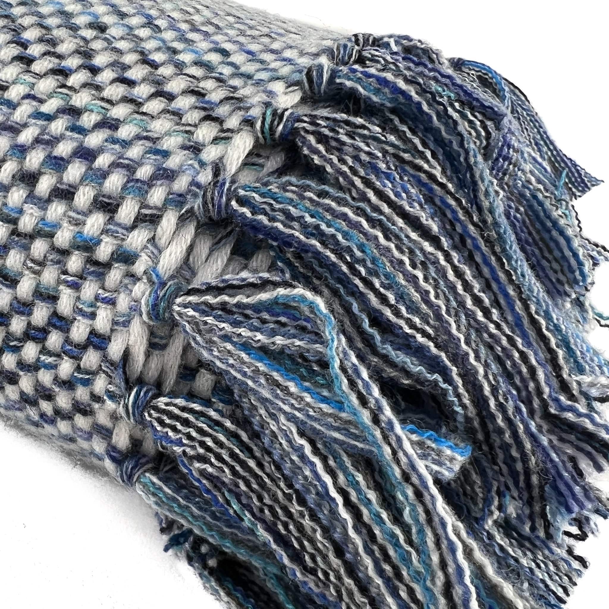 Close-up detail of an Italian made Cashmere throw with a white weft and different shades of blue warp, folded on a white background with handknotted multiple shades of blue tassels showing in front