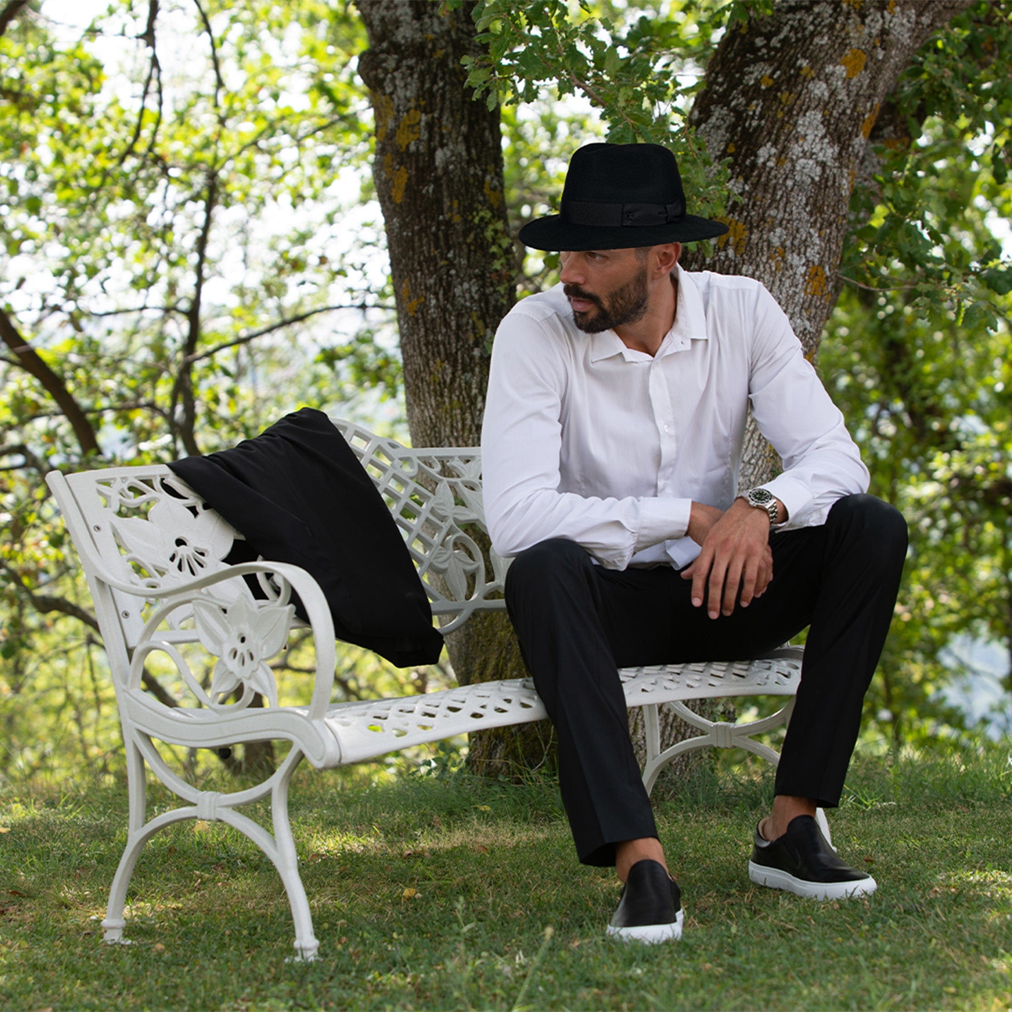 Man sitting on white bench wearing a black Virgin wool light felt fedora hat with black gross grain ribbon and bow, with iconic black crafted society scissor on the left side. 100% virgin wool, water repellent and crushable. 
